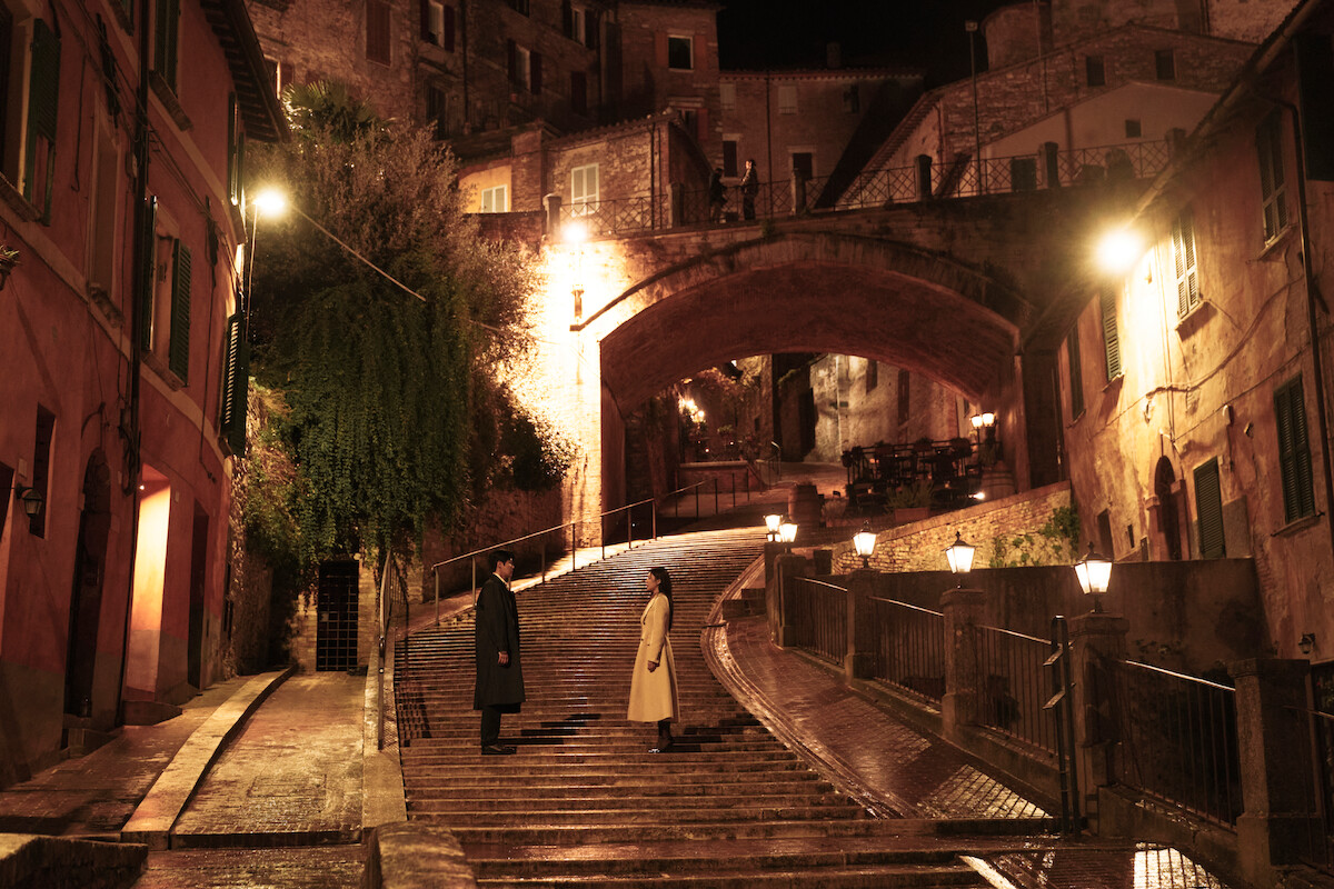 Two people stand facing each other on a wet, illuminated stone staircase at night in an old European city, surrounded by historic buildings, greenery, and street lamps with a large archway above.