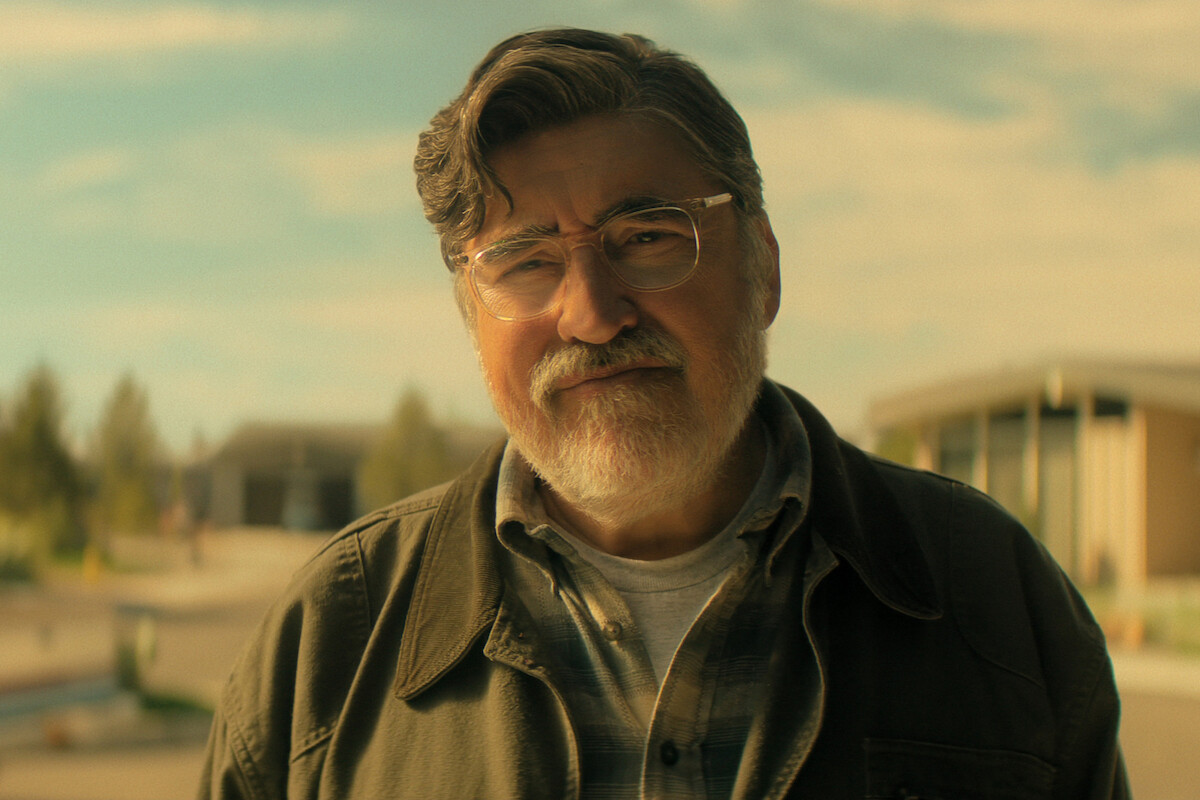 Alfred Molina as Sam, with glasses and gray hair standing outdoors, wearing a jacket and plaid shirt on a sunny day, with modern buildings and trees blurred in the background.