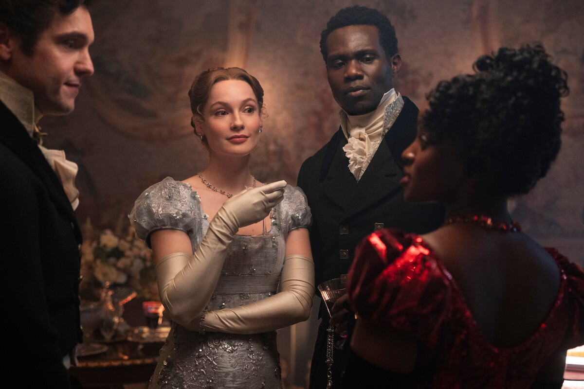 Four people in elegant Regency-era costumes converse in a dimly lit, ornately decorated room, suggesting a historical or period drama setting, with one woman in a silver gown and another in red.