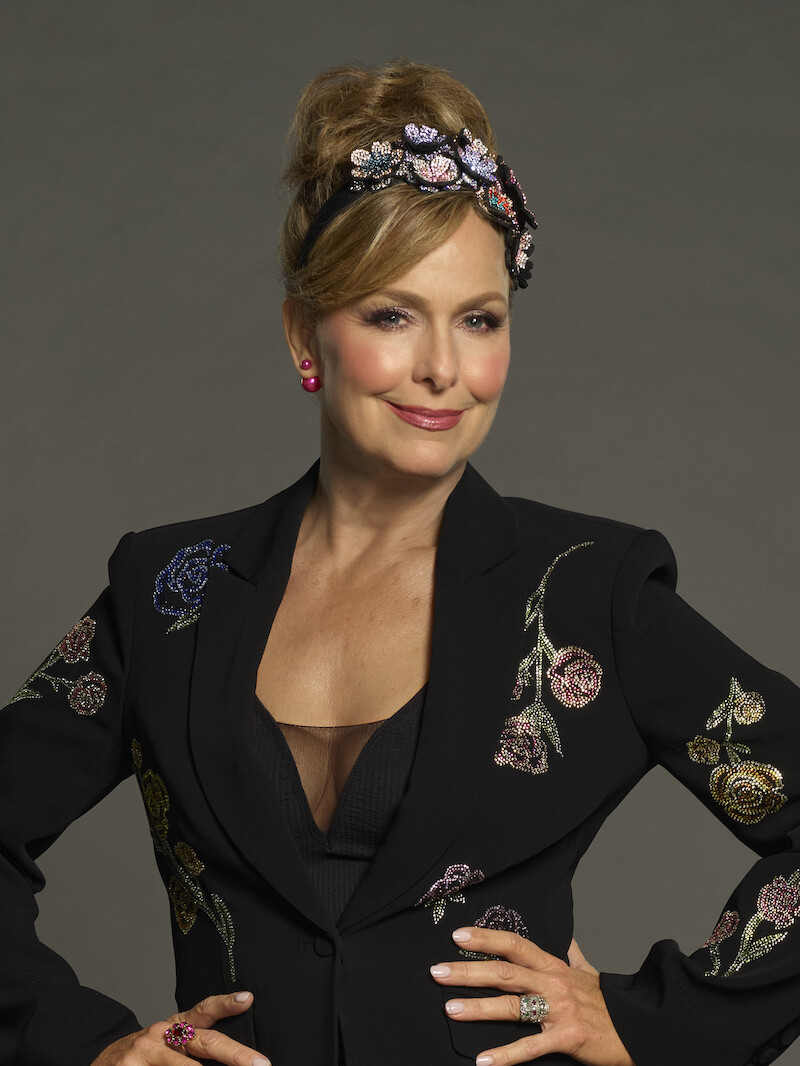 Mulher usando um blazer preto com flores bordadas e uma tiara com pedras brilhantes faz uma pose confiante com as mãos nos quadris, usando brincos rosas e anéis, em um estúdio com plano de fundo neutro e liso.