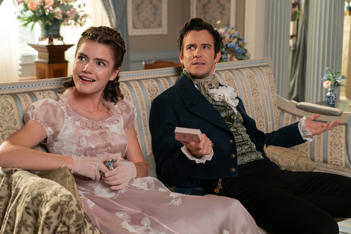 A young woman in a pink Victorian dress and a man in a dark suit sit on a patterned sofa in an elegant, flower-filled room, appearing to converse and relax together.