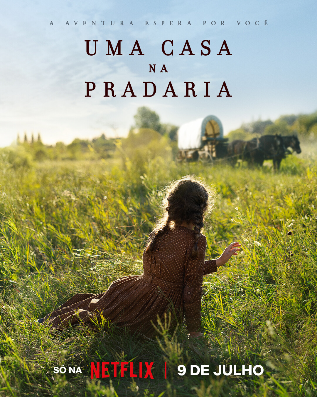 Garota sentada em um gramado observando uma carroça coberta e cavalos em uma pradaria ensolarada. Imagem promovendo ‘Uma Casa na Pradaria’, disponível na Netflix em 9 de julho.