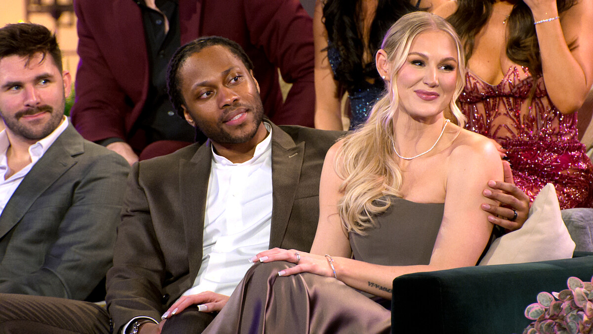 Three well-dressed people sitting closely on a couch at a formal indoor event, likely a television show or gathering, with an elegant, festive background and audience members dressed in evening attire visible behind them.