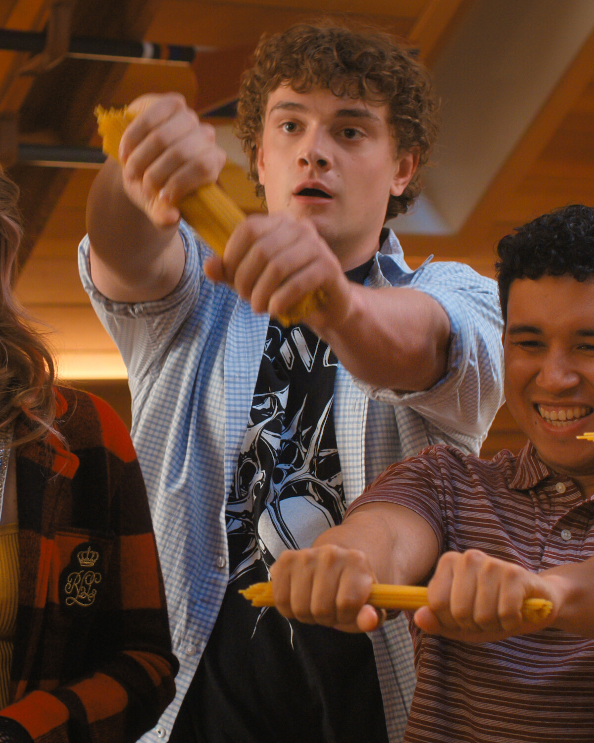 Two young men indoors enthusiastically break uncooked spaghetti in half, surrounded by friends, in a brightly lit room with a wooden ceiling, capturing a playful and energetic moment.