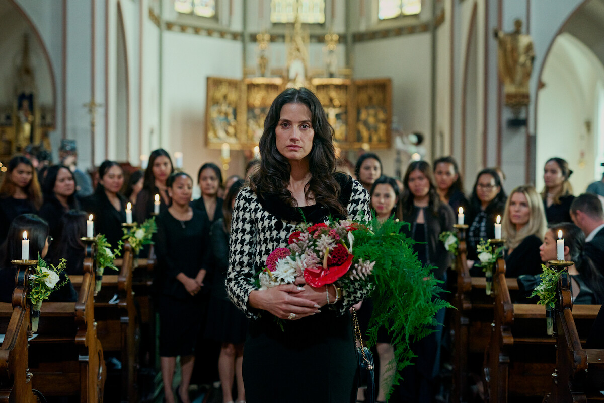In an image from the series ‘Secrets We Keep,’ Danica Curcic as Katarina holds flowers while walking down a church aisle as a large group of women dressed in black congregate behind her.
