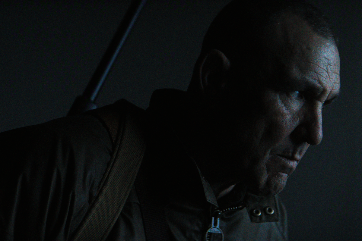 A man with a serious expression is in a dimly lit setting, wearing a brown jacket and carrying a strap over his shoulder, suggesting a tense or dramatic atmosphere.