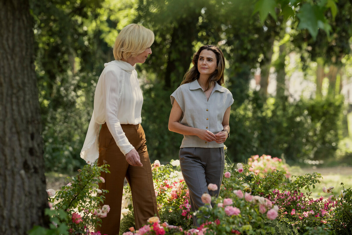 Two women having a conversation while walking through a blooming garden surrounded by green trees and colorful flowers on a sunny day.