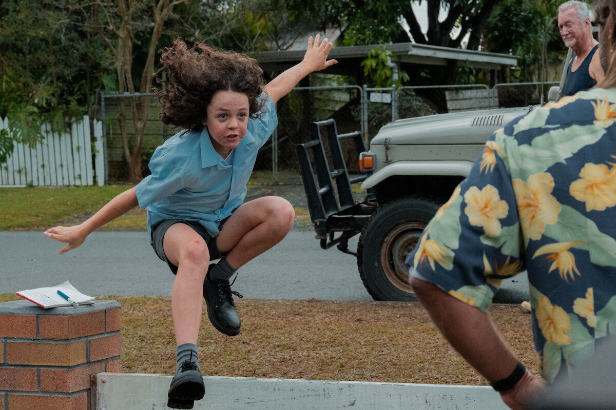 Felix Cameron as Eli Bell jumps over a fence toward Bryan Brown as Slim Halliday in a still image from the series ‘Boy Swallows Universe.’
