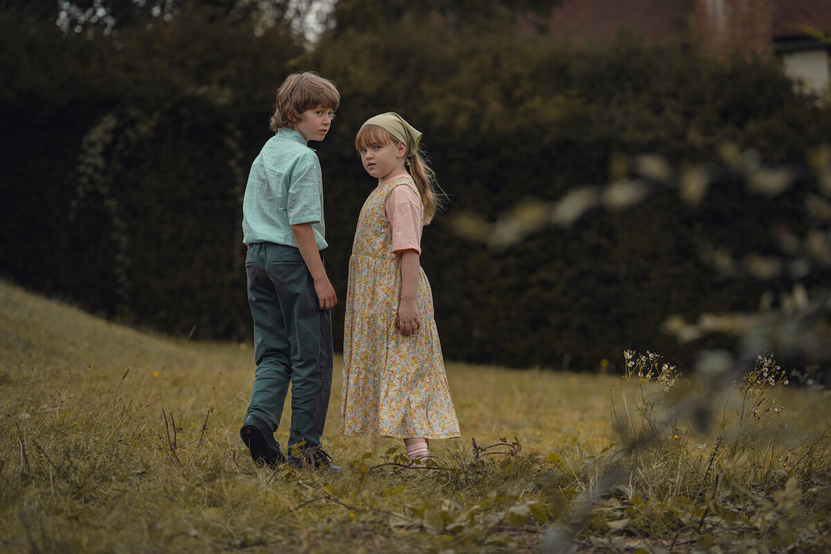 Two children stand close together in a grassy field, surrounded by greenery and trees, with an overcast or moody atmosphere, both looking back toward the camera with serious expressions.