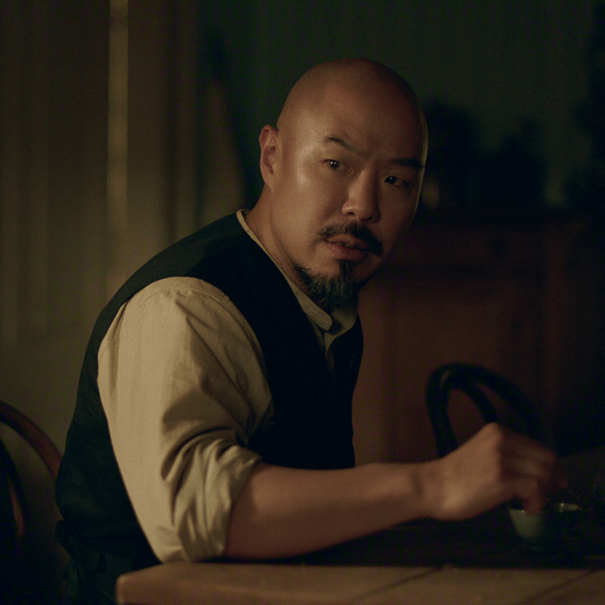Hoon Lee as Lee with a shaved head and goatee sits at a wooden table in a dimly lit room, wearing a beige shirt and dark vest, looking to the side with a thoughtful expression.Hoon Lee as Lee with a shaved head and goatee sits at a wooden table in a dimly lit room, wearing a beige shirt and dark vest, looking to the side with a thoughtful expression.