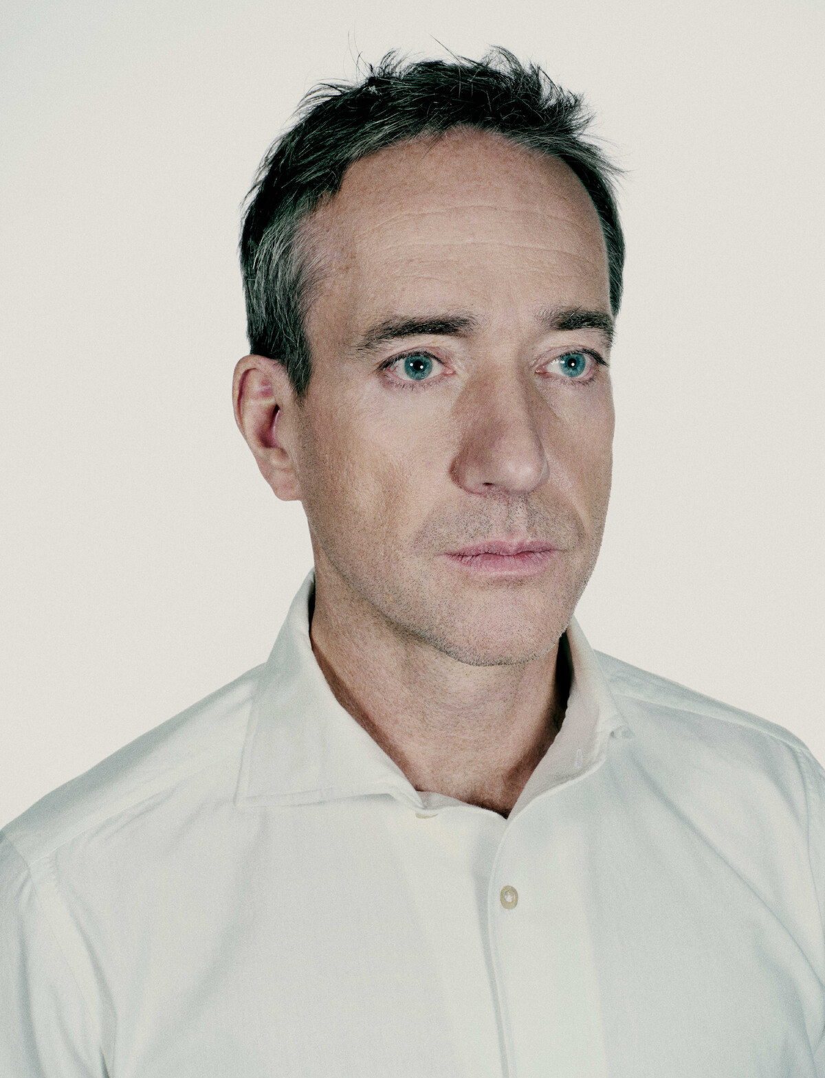 Man with short hair in a white shirt stands against a plain light background, looking slightly away from the camera with a serious, contemplative expression. The mood is subdued and introspective.