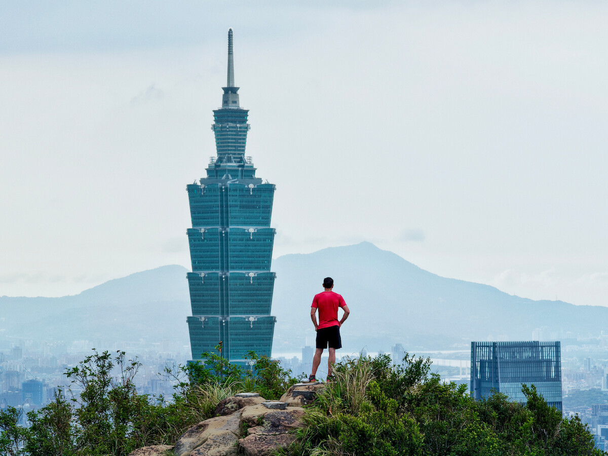 Alex Honnold in athletic clothing stands on a rocky hilltop, overlooking the city skyline with the Taipei 101 skyscraper and distant mountains in the background under a cloudy sky.