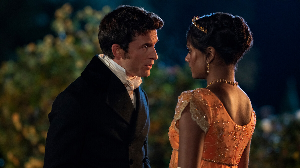 A man and woman in historical clothing stand close together outdoors at night, surrounded by greenery, appearing to have an intense conversation.