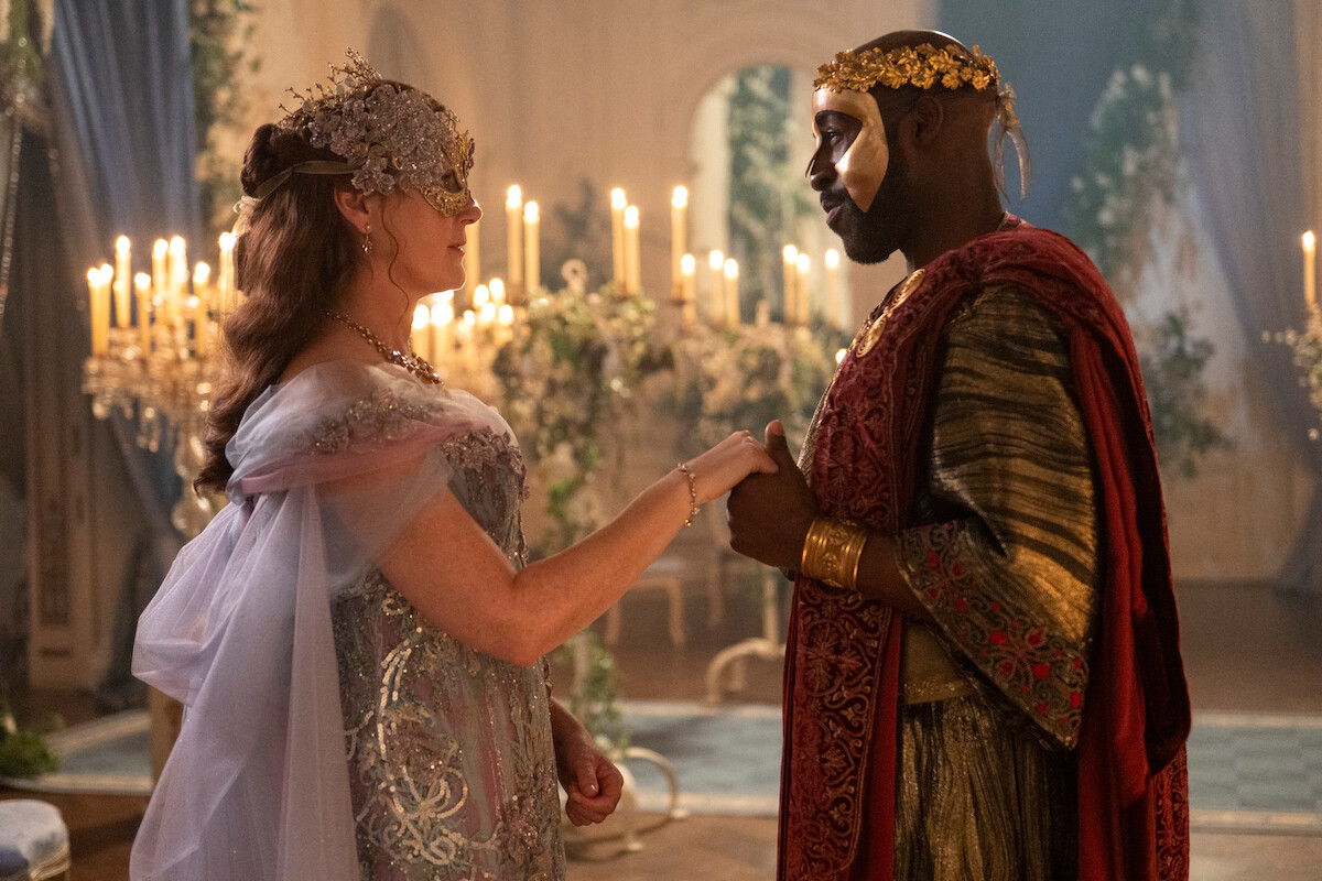 Two people in elaborate masquerade costumes hold hands in a candlelit, ornate room with draped curtains and floral decor, creating a regal and romantic atmosphere.