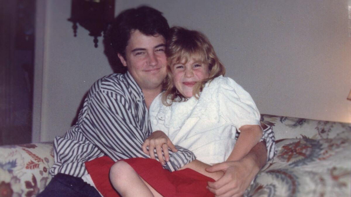 Man and young girl smiling and sitting on a floral-patterned couch indoors, with warm lighting and casual attire, creating a cozy, affectionate family moment.