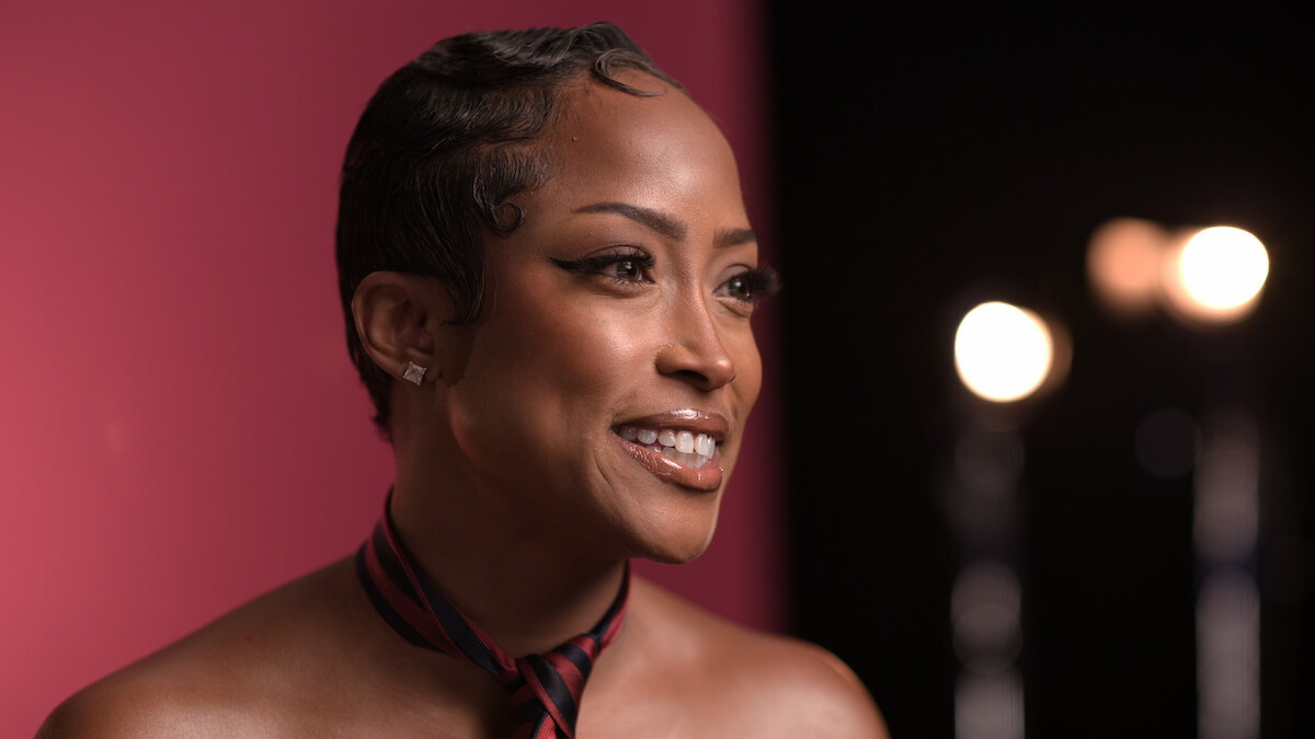 Smiling woman with short hair and a striped necktie poses in a studio with a pink and black background and blurred stage lights.