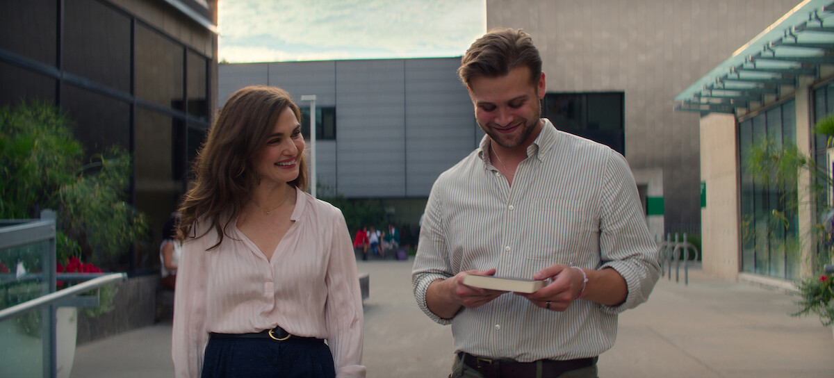 Rachel Weisz as M and Leo Woodall as Vladimir walk outdoors in a modern urban setting. The woman smiles while the man looks down at a book or notebook in his hands. They appear relaxed and friendly on a paved pathway near buildings and greenery.