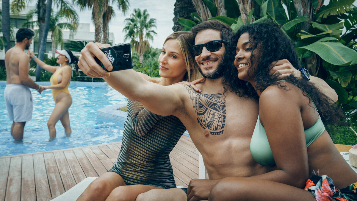 A man with a tattoo takes a selfie with two women by a swimming pool
