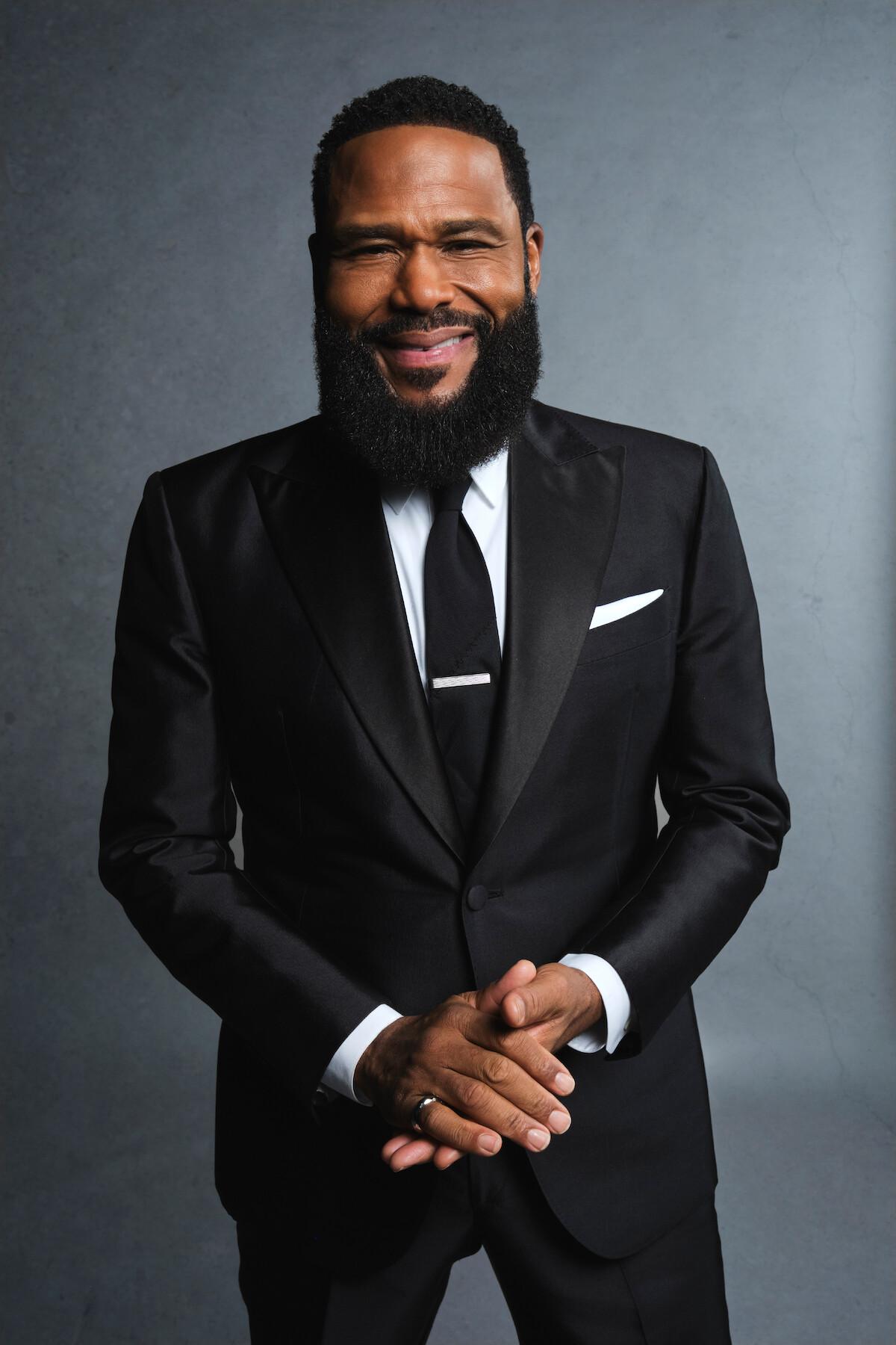 A confident man with a full beard smiles warmly while wearing a black tuxedo, white shirt, and tie. He stands against a neutral gray background, exuding elegance and a positive, approachable mood.