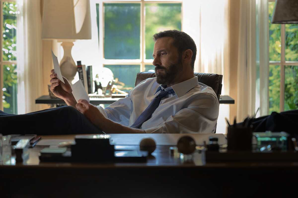 Man in dress shirt and tie sits at a desk in a sunlit office, reading papers with feet up. Desk has pens, papers, and lamps. Large windows and trees outside create a bright, contemplative setting.