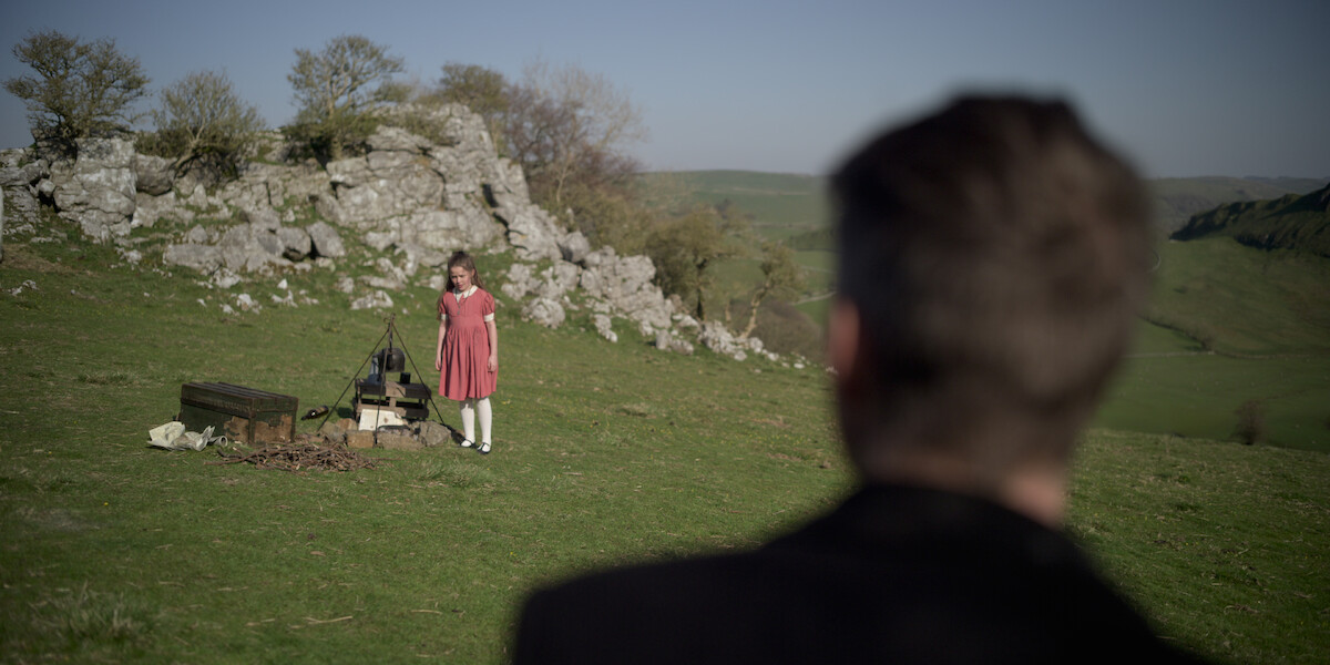 A man looks over to a small girl in a field who is in a pink dress and near a fire.