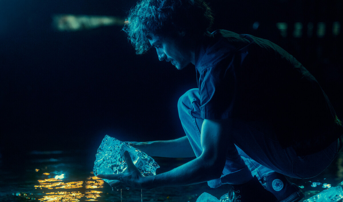 Person crouched by water at night, illuminated by blue light, holding a large ice-like object, with reflections on the surface, suggesting a calm outdoor setting.