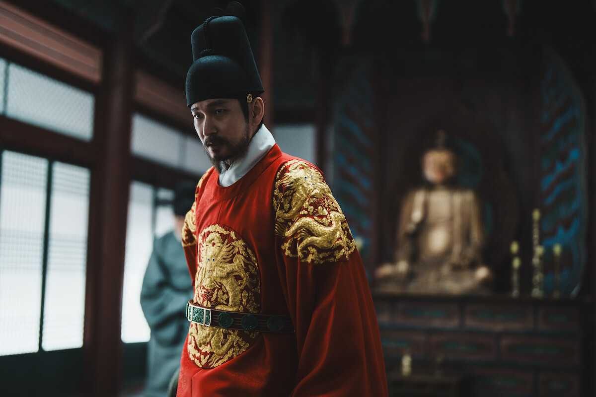 Man dressed in traditional royal Asian attire stands in an ornate room with wooden details and a large golden Buddha statue in the background, suggesting a historical or ceremonial setting.Man dressed in traditional royal Asian attire stands in an ornate room with wooden details and a large golden Buddha statue in the background, suggesting a historical or ceremonial setting.