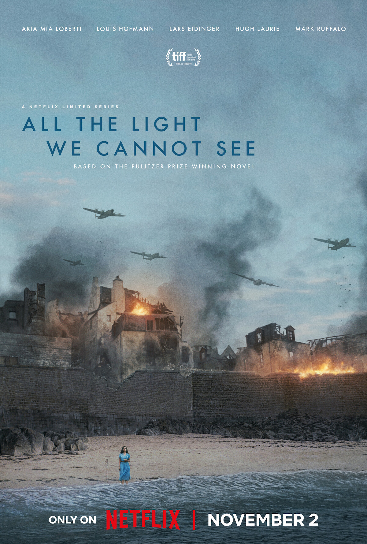 ‘All the Light We Cannot See’ key art - a woman in a blue dress standing on a beach in front of a burning town. 