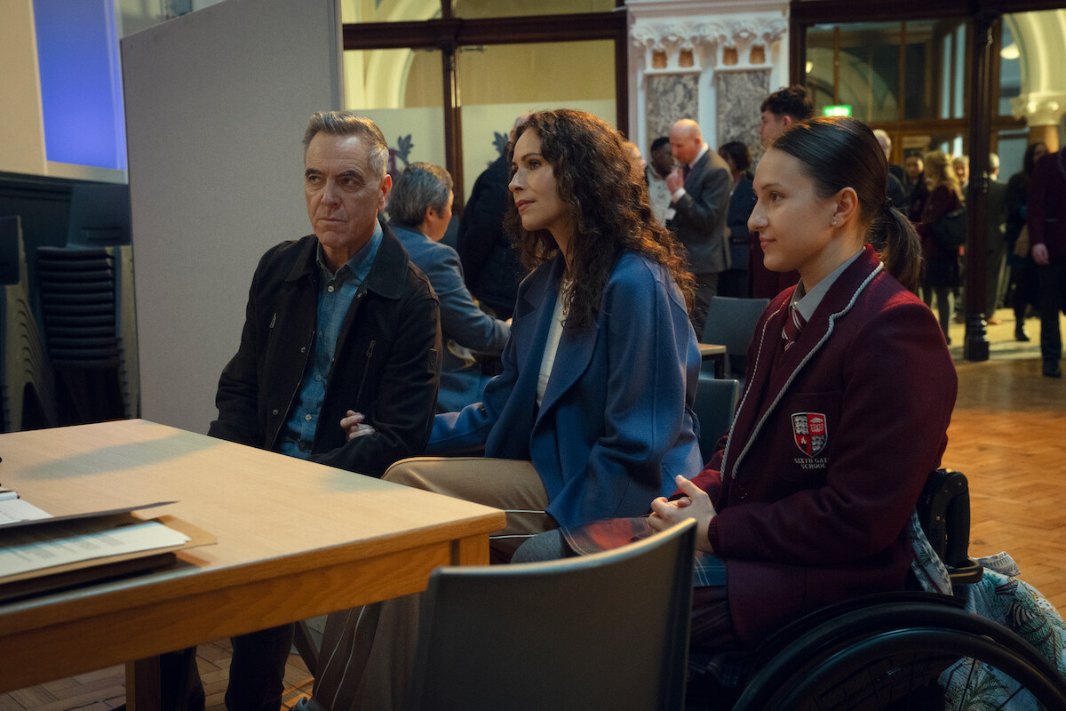 Three people, including a man, a woman, and a girl in a wheelchair wearing a school uniform, sit at a wooden table in a large hall with people in the background, giving a serious, contemplative mood.