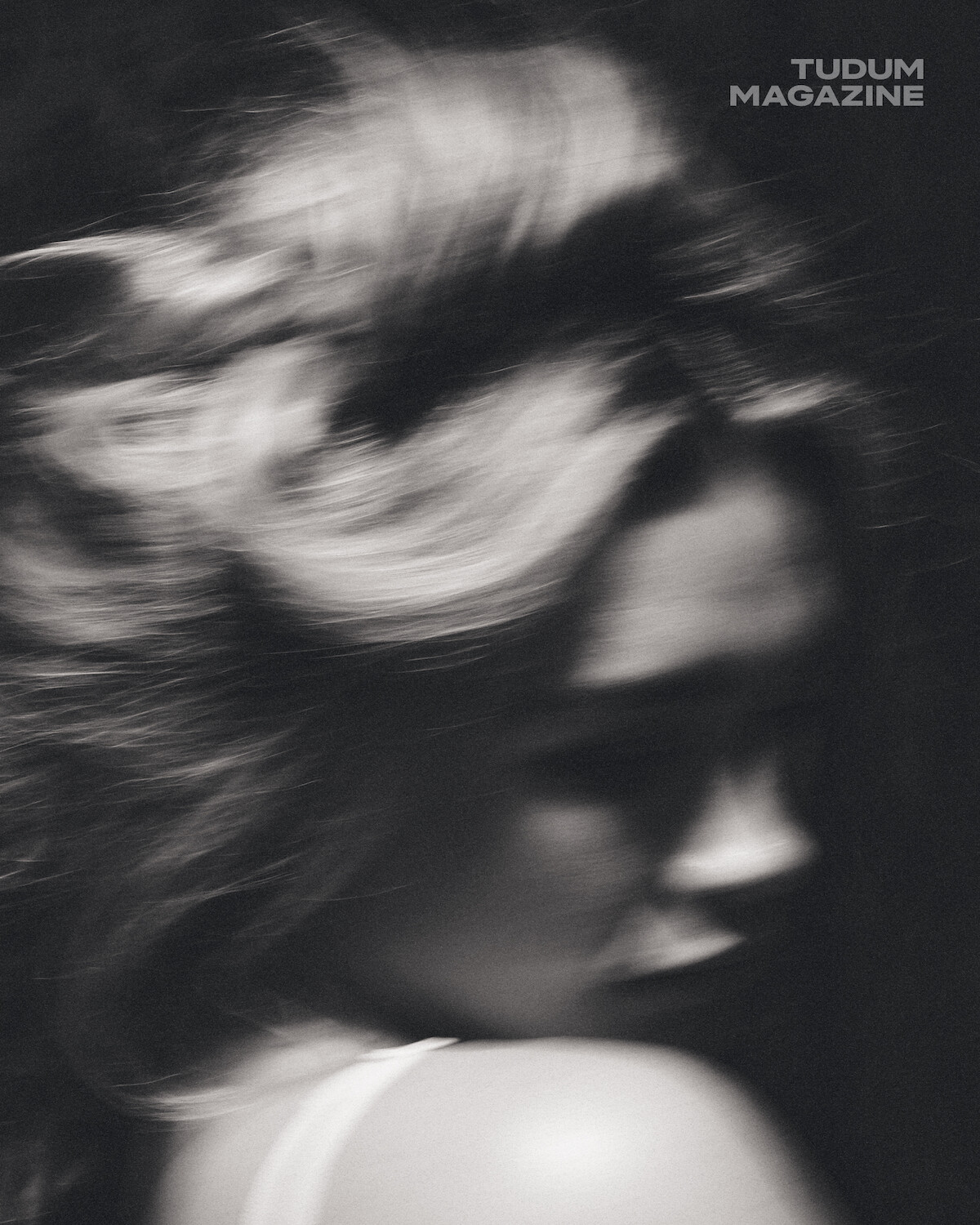 Blurry black-and-white photo of a person with long hair turned away, soft lighting, moody atmosphere; "TUDUM MAGAZINE" text in the upper right corner.