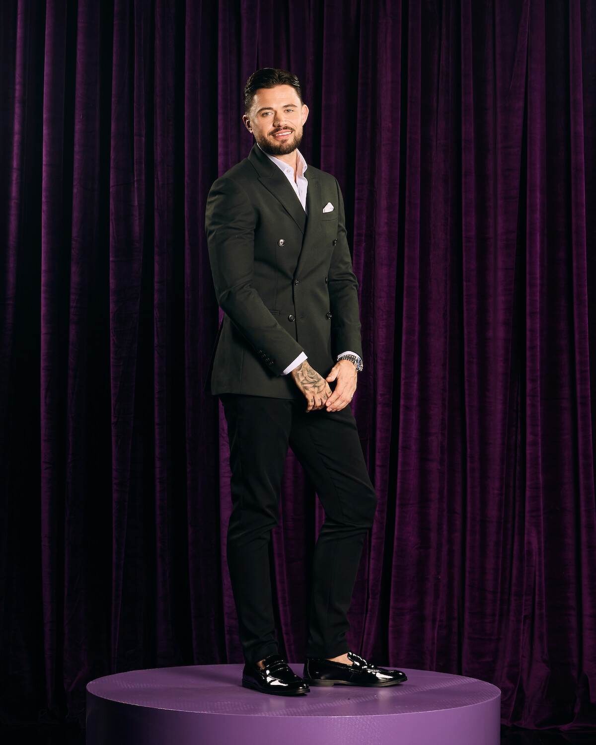 A well-dressed man with tattoos stands on a round purple platform in front of dark purple curtains, wearing a suit jacket, dress shirt, and loafers in a formal indoor setting.