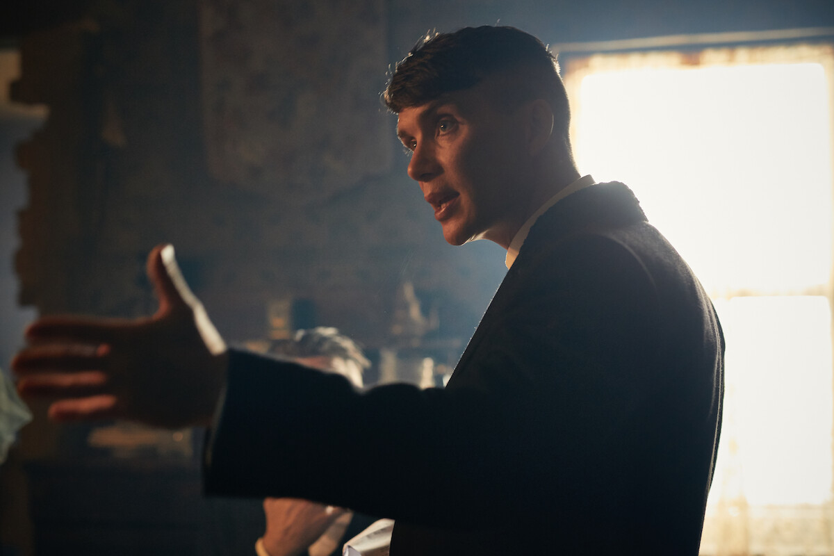 Man in a suit gesturing with his hand, standing in a dimly lit room with a large sunlit window and rustic decor in the background.