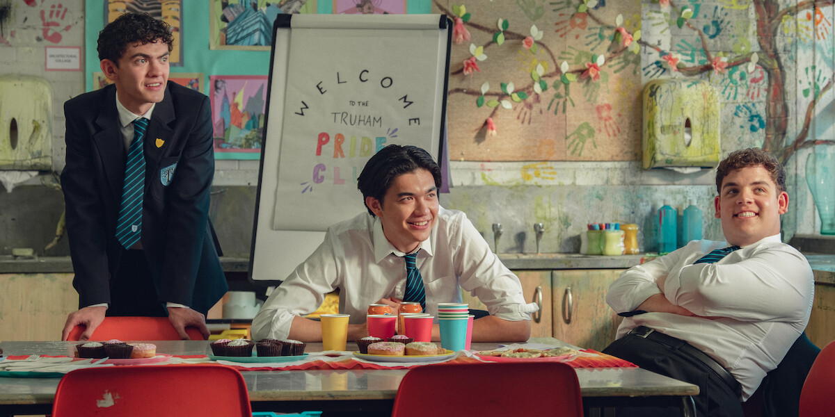 Three students in school uniforms sit and smile around a table with snacks and drinks in a colorful classroom decorated with children's artwork and a "Welcome to the Truham Pride Club" sign in the background.