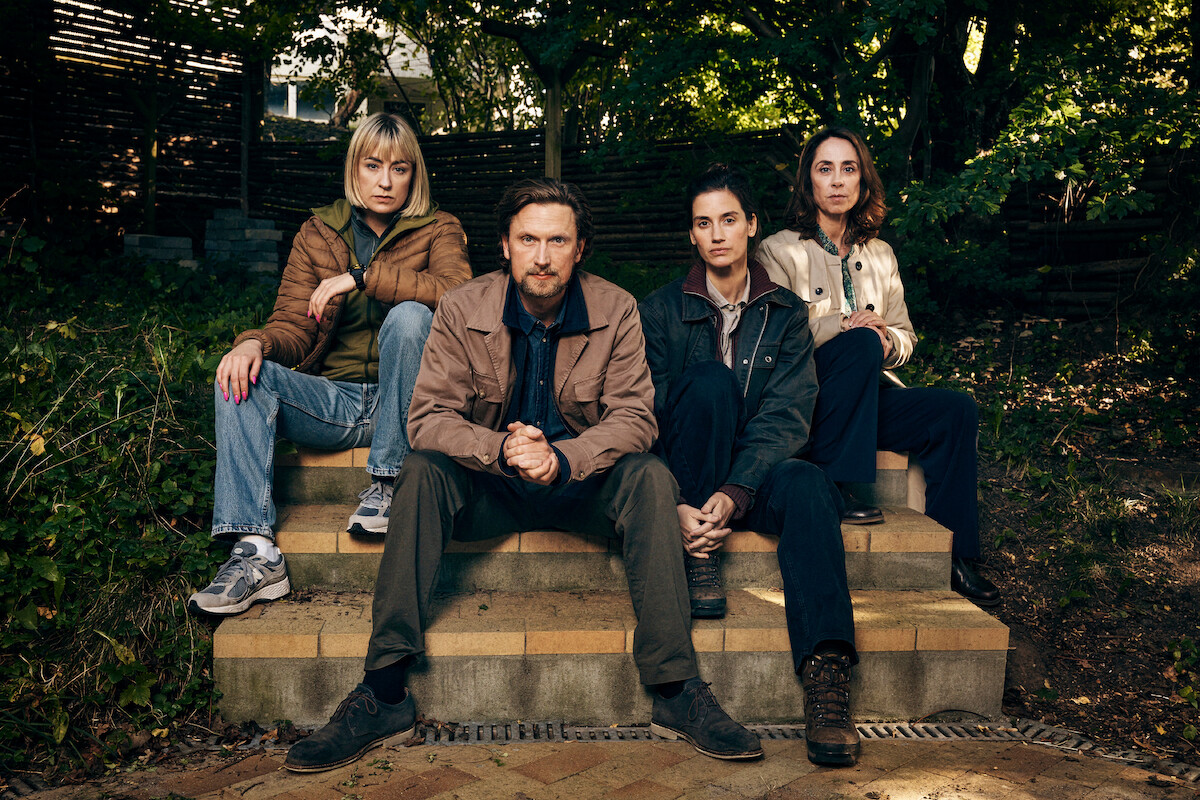 Four people sit on outdoor steps surrounded by greenery and wooden fencing, wearing casual jackets and pants, looking directly at the camera with serious expressions in a shaded garden setting.