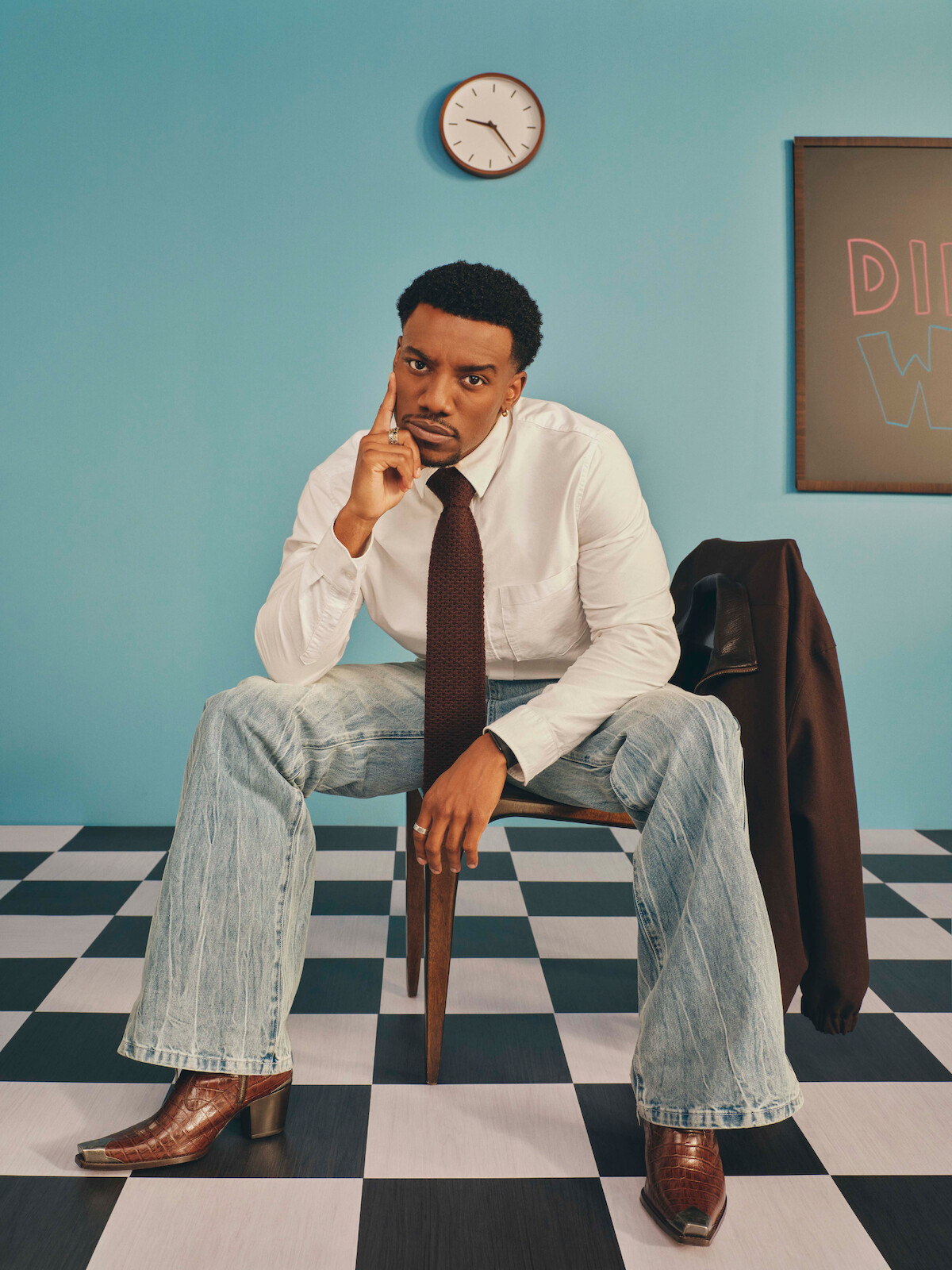 Man in a white shirt and tie sitting on a chair in a room with blue walls, checkered floor, a clock above, and a jacket draped on the chair; setting appears retro and contemplative.