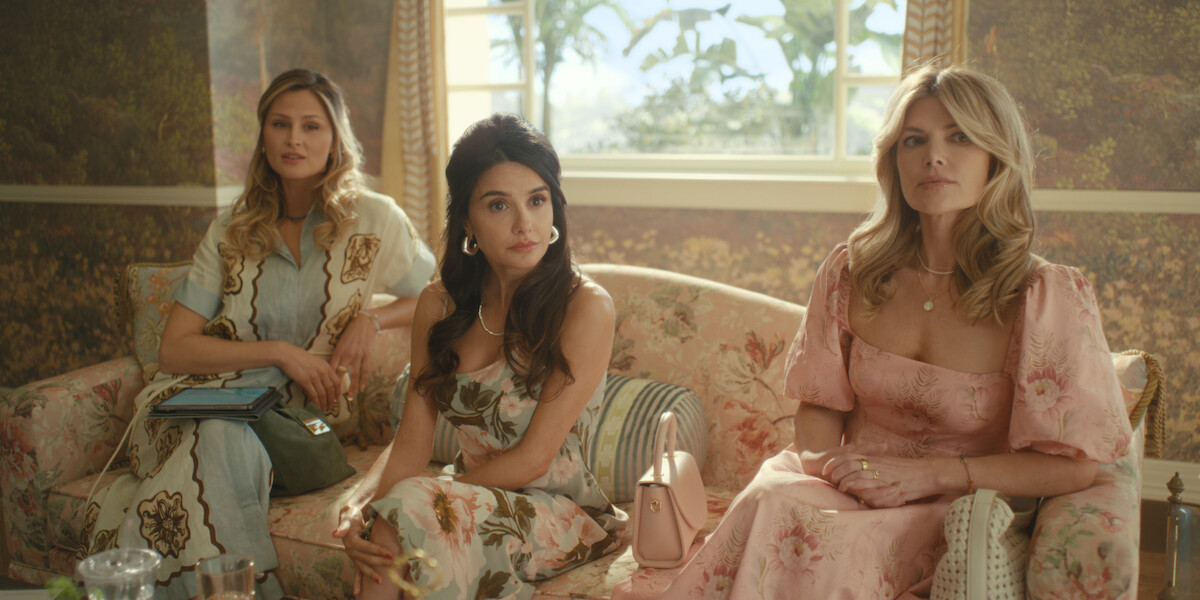 Three women in elegant floral dresses sit on a patterned sofa in a well-lit, stylish living room with large windows and decorative wallpaper, creating a sophisticated, relaxed atmosphere.