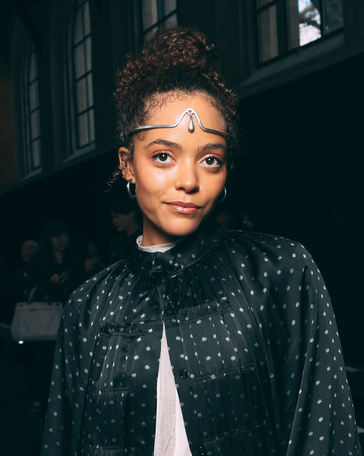 Woman with curly hair in a polka-dot top and silver headpiece, standing indoors at an event or gathering with large windows and people in the background.