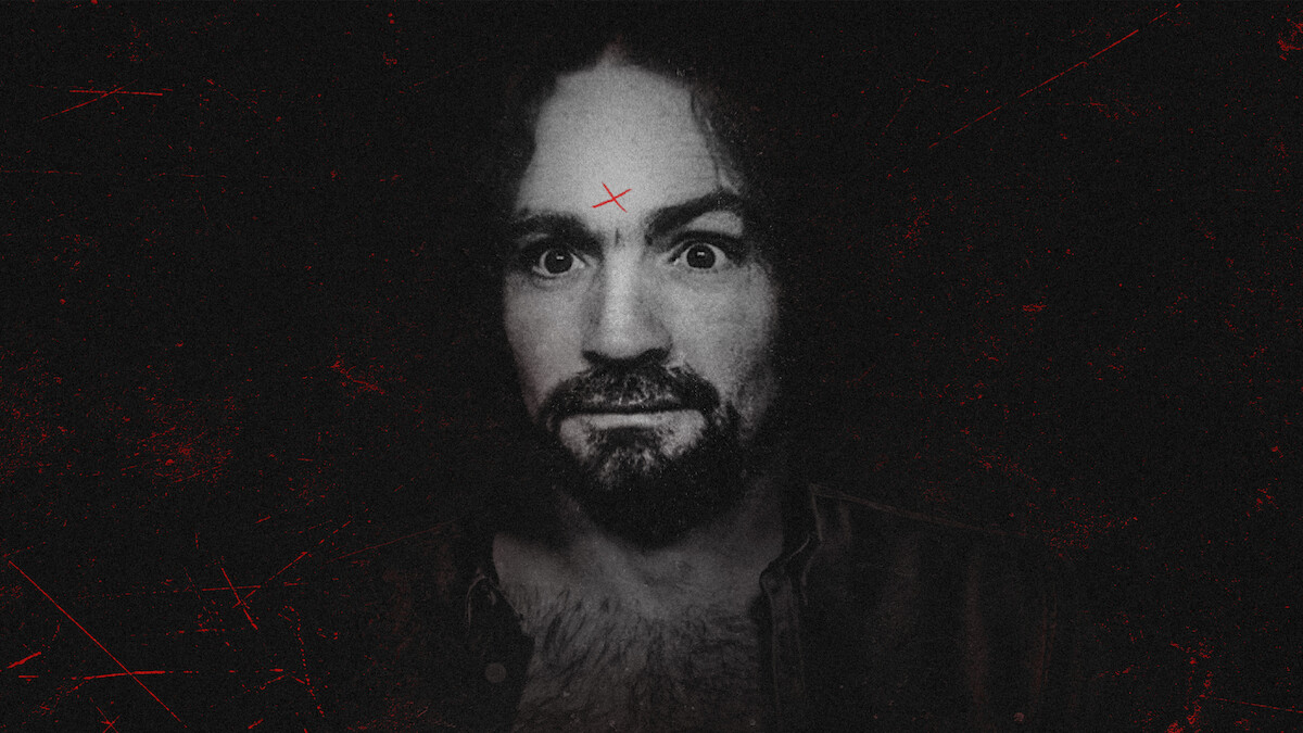 A man with curly hair and a beard stares intensely at the camera, set against a dark, gritty background. He has a red X marked on his forehead and visible chest hair, wearing an open shirt. The overall mood is eerie and dramatic.