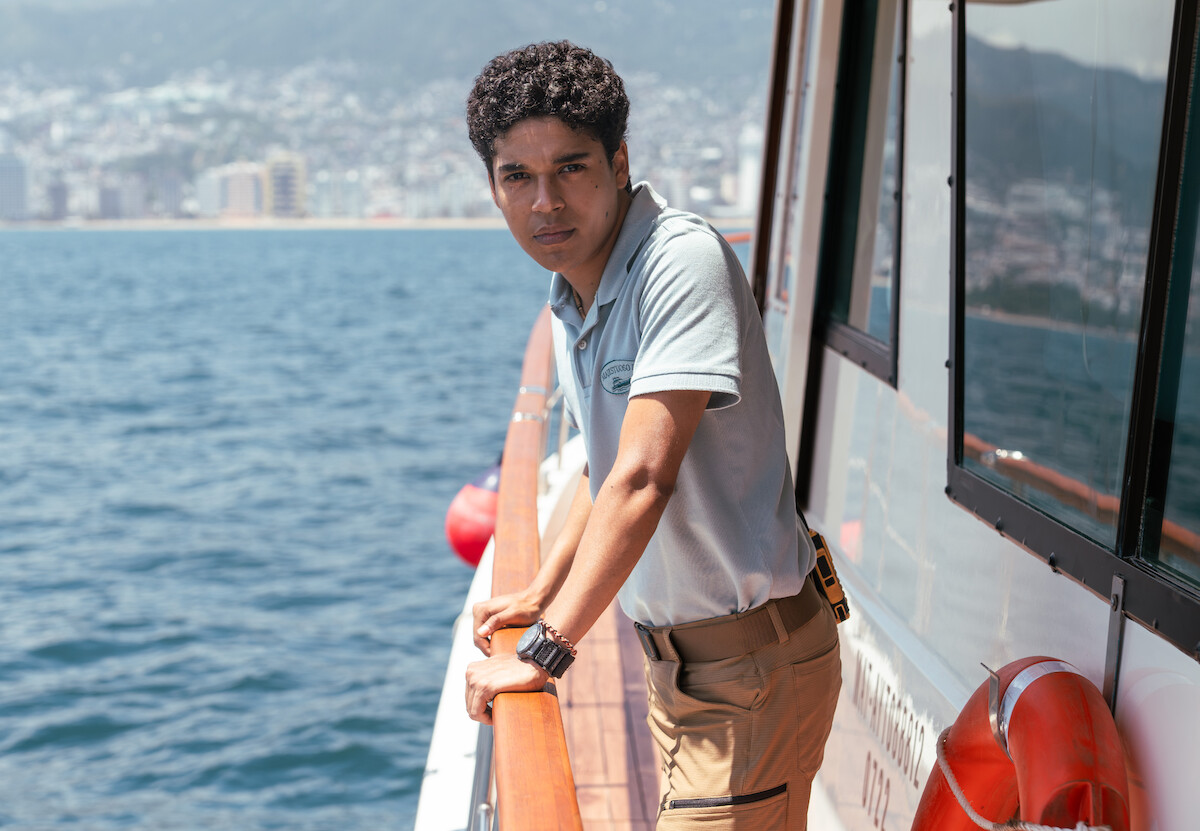 Um homem de cabelos curtos e encaracolado, vestindo camisa azul e uma bermuda bege, apoia as mãos na grade de um barco. Ao fundo é possível ver o mar e, no limite do horizonte, uma cidade fora de foco
