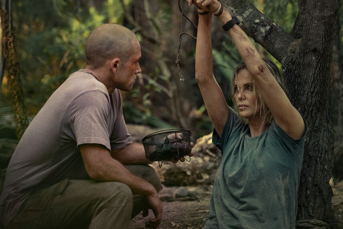 A man kneels in a forest facing a woman who is tied to a tree with her hands bound above her head. The woman has visible bruises and looks distressed. The setting is outdoors with trees and natural light.