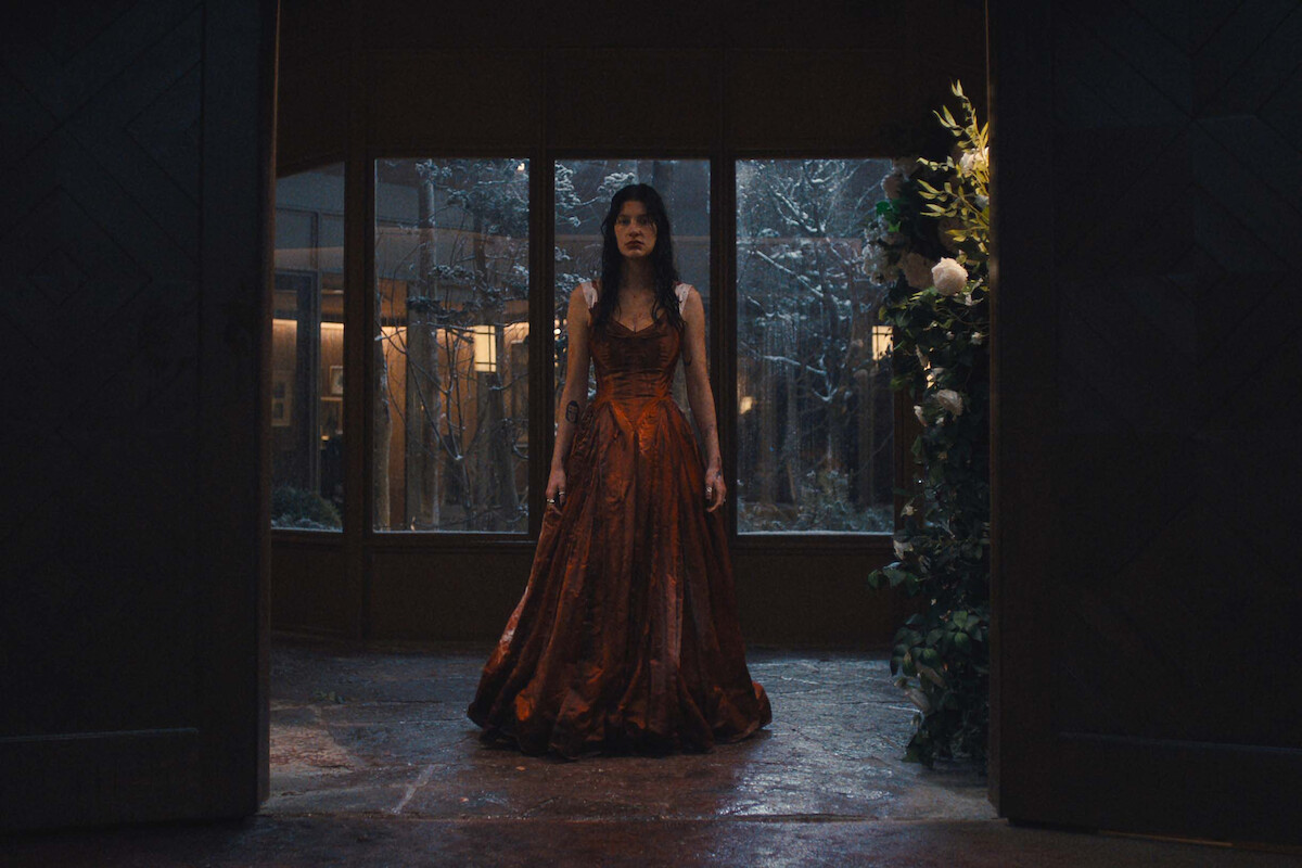 A woman in a red gown stands in a dimly lit, elegant room with stone floors, large windows showing a snowy scene outside, and a floral arrangement to the right.