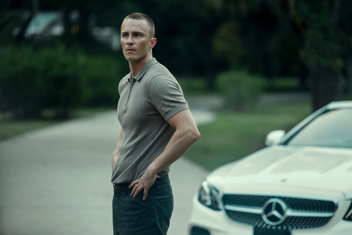 A man stands outdoors on a driveway next to a white Mercedes-Benz car, surrounded by greenery on a cloudy day.