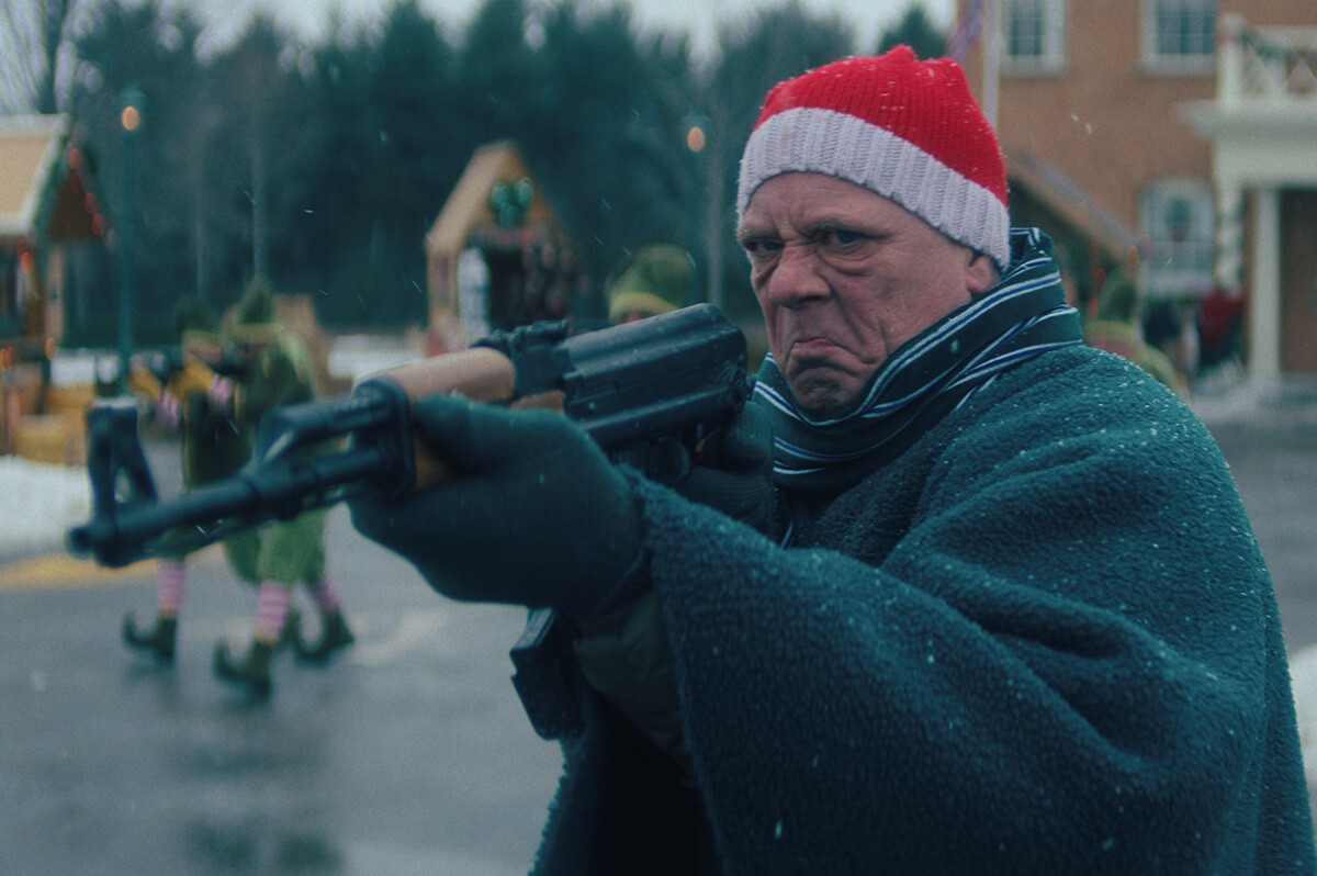 A man with a Santa Claus beanie and cape pointing a rifle with a disgruntled facial expression.