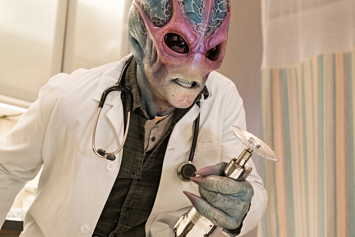 Alien in a lab coat with a stethoscope holds a futuristic medical device in a clinical or hospital setting, suggesting a sci-fi or medical experiment scene.