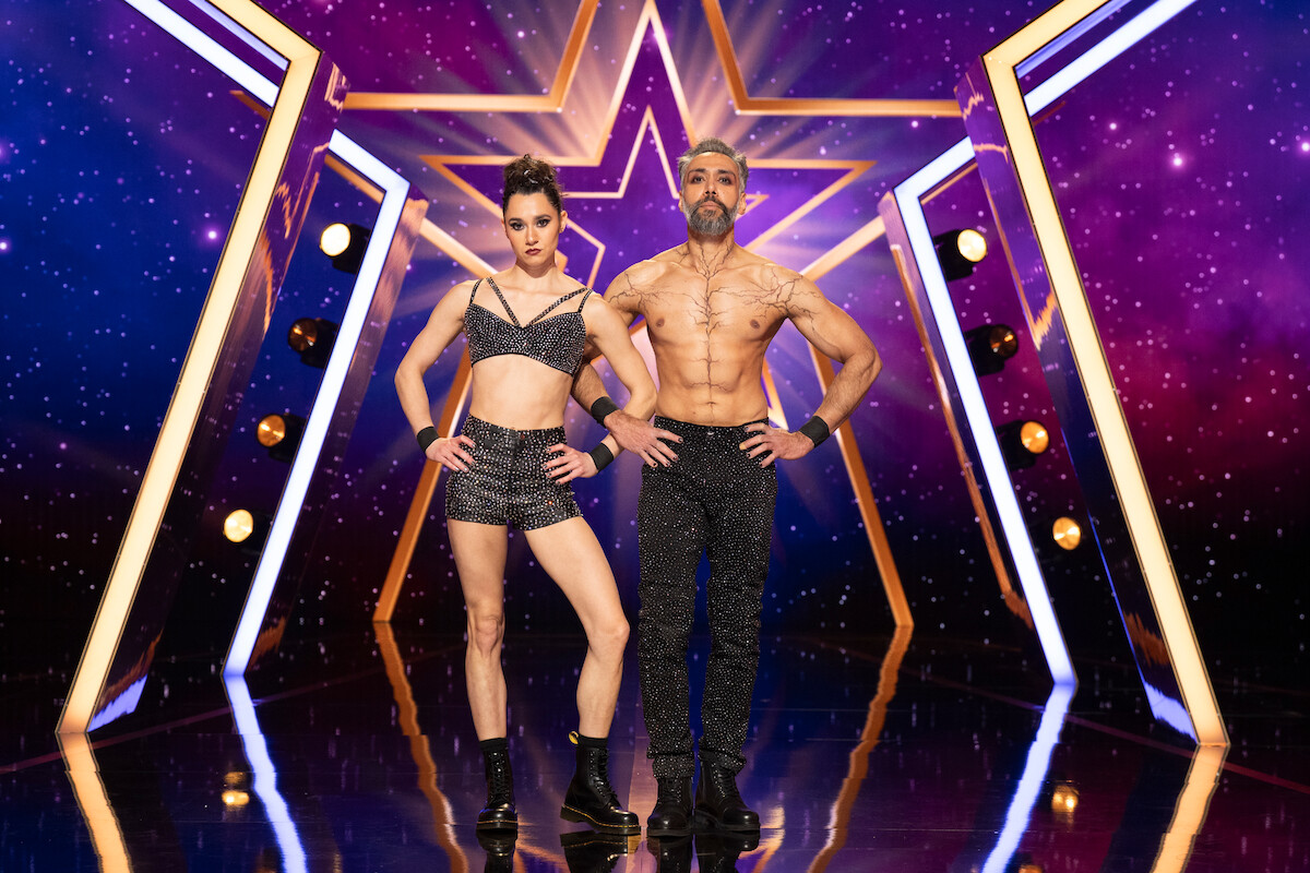 Duo Vespertilio in sparkly performance outfits stand confidently on a shiny stage with a star-shaped neon backdrop and purple starry lights, ready for a talent show performance.