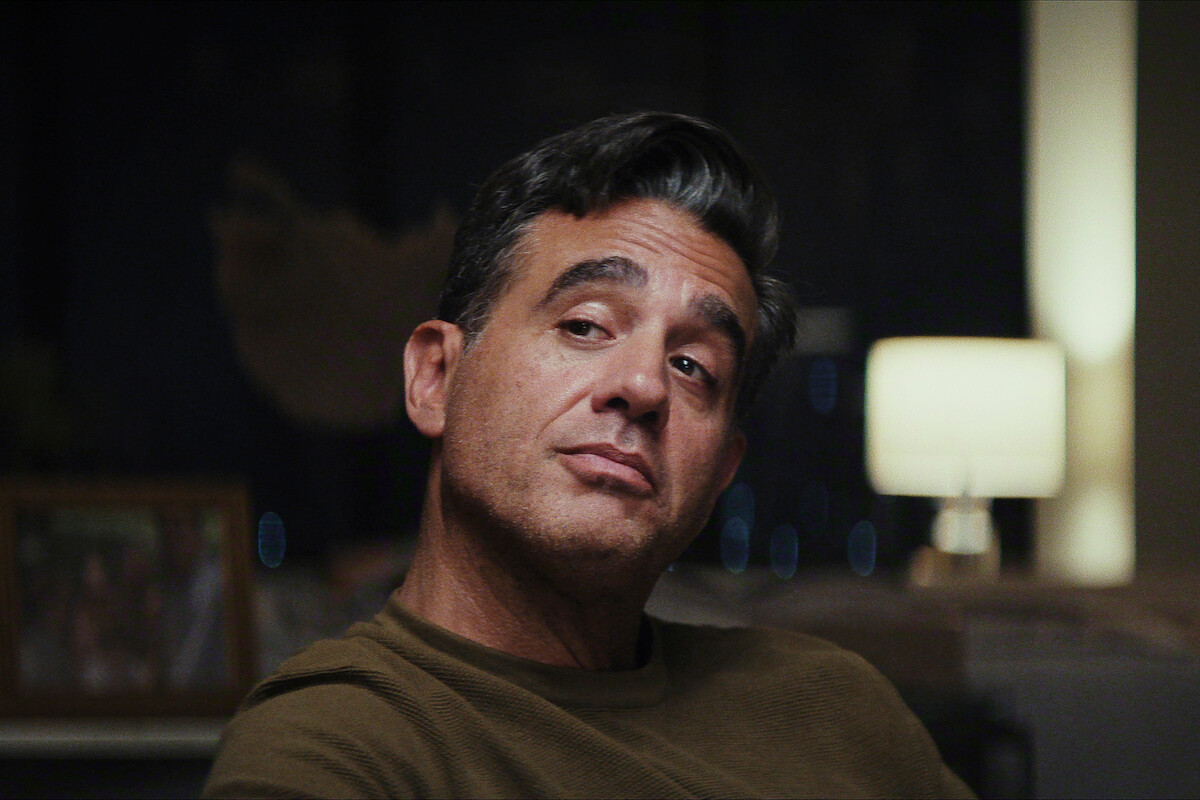 Man with short dark hair wearing a brown shirt sits indoors, looking to the side with a thoughtful expression. The background includes a softly lit lamp and a blurred photo frame, creating a cozy, dimly lit atmosphere.
