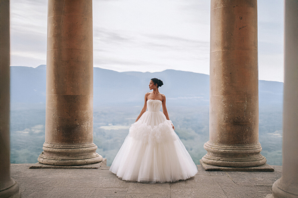 Bride in elegant white gown stands between stone columns on a terrace, overlooking misty mountains and valley, creating a serene and romantic atmosphere.