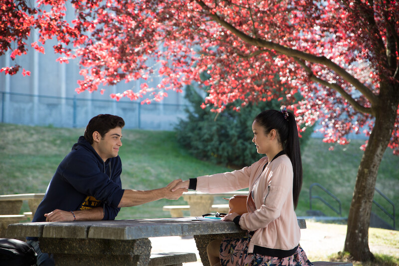 Noah Centineo as Peter and Lana Condor as Lara Jean sitting at a table shaking hands.