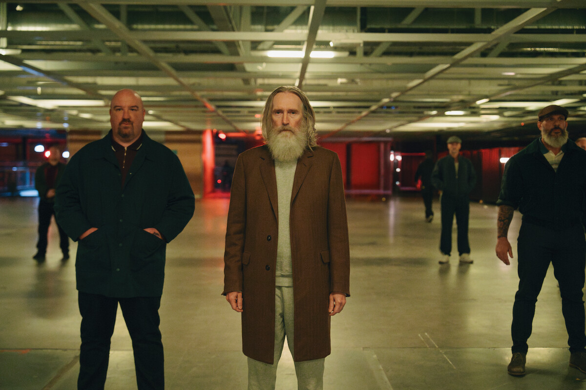 A group of men, including an older man with a long white beard in a brown coat, stand in a dimly lit, industrial-style room with exposed ceiling beams and concrete floors.