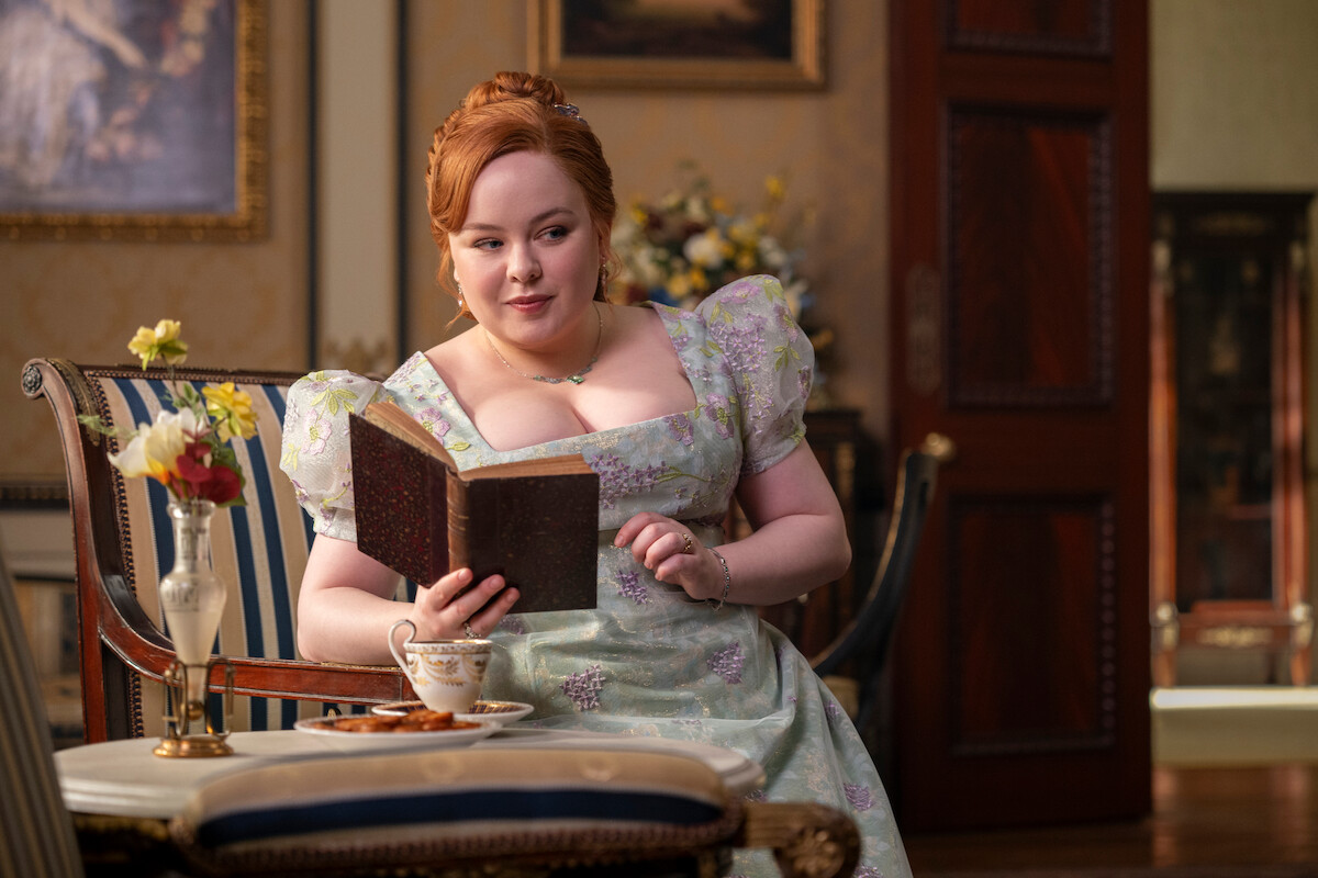 Woman in a floral Regency-era dress sits on a striped chair, reading a book in an elegant, ornately decorated room. There is a table with tea and flowers beside her, and framed paintings hang on the wall in the background.